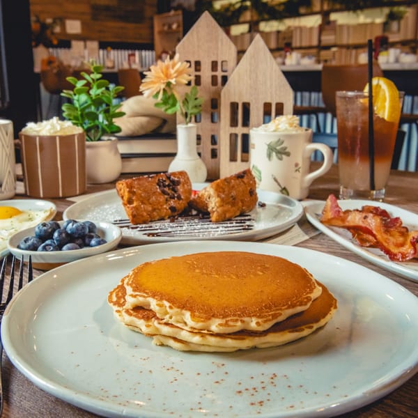Breakfast spread with pancakes, blueberries, bacon, pastries, and drinks on a wooden table.
