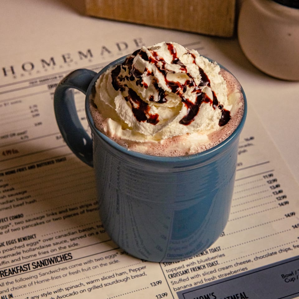 A blue mug of hot chocolate with whipped cream and chocolate drizzle sits on a menu.