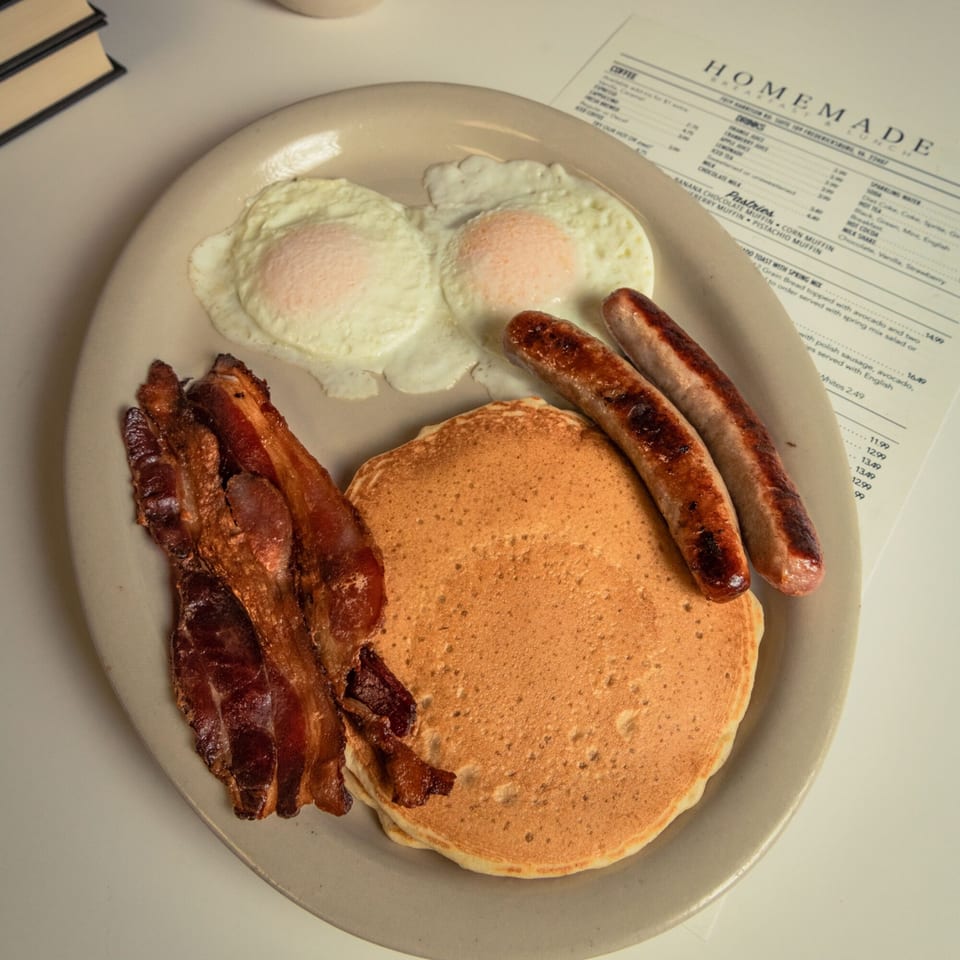 A breakfast plate with pancakes, bacon, sausages, and two fried eggs, next to a menu.