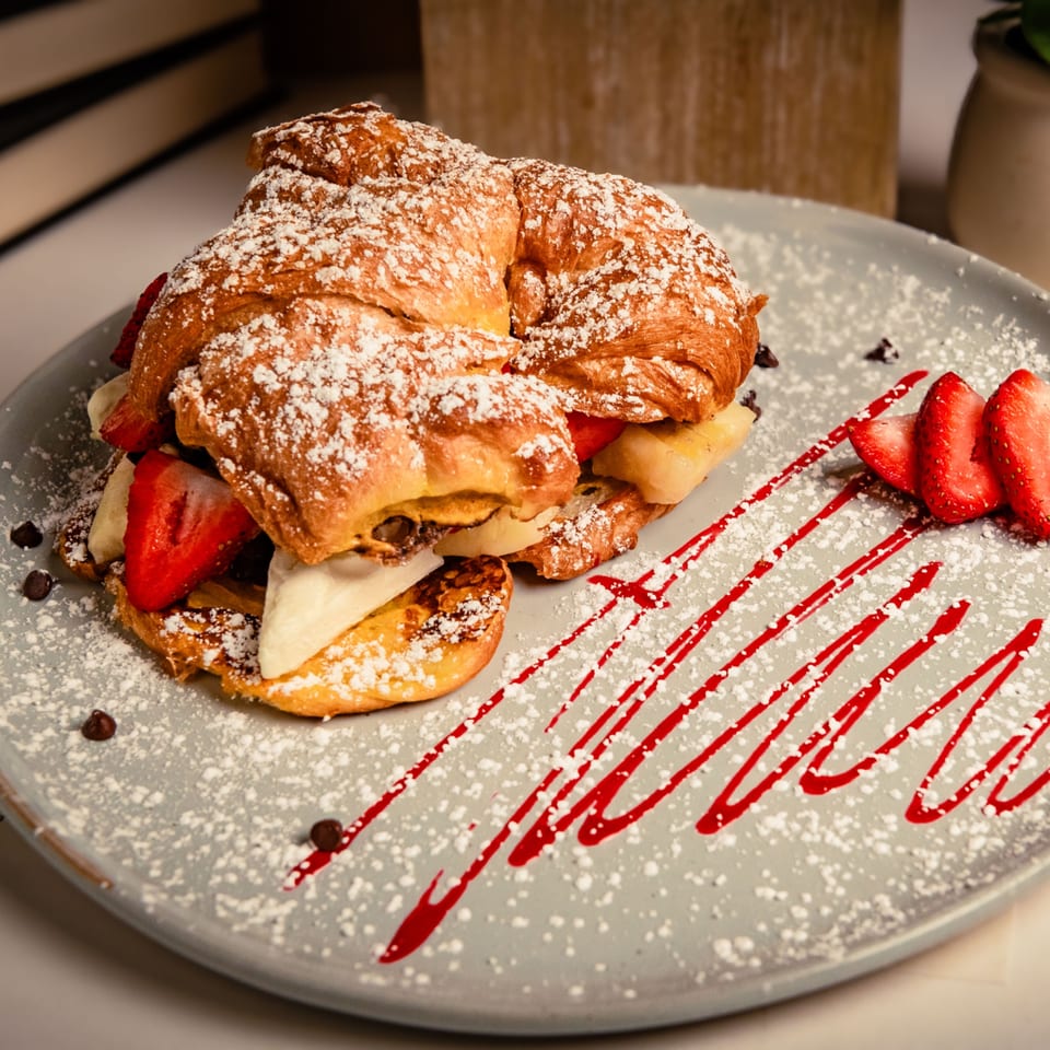 Powdered sugar-dusted croissant filled with strawberries, bananas, and cream, drizzled with raspberry sauce.