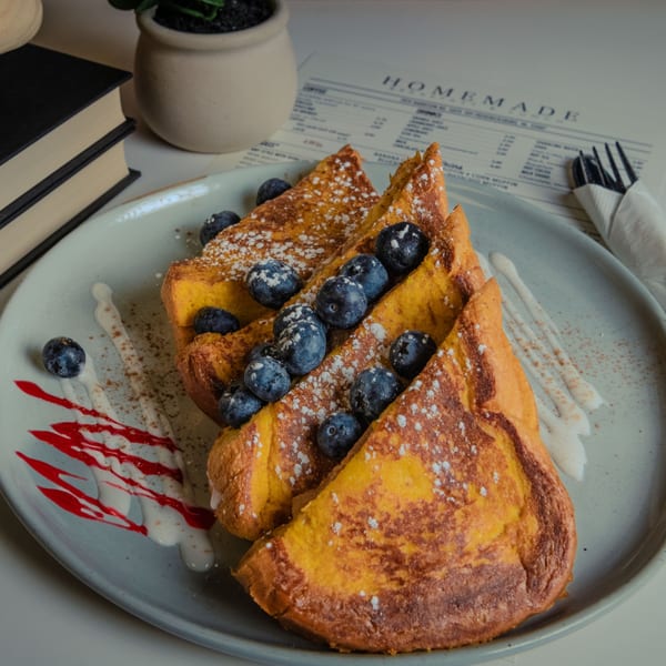 Golden French toast slices topped with blueberries, powdered sugar, and white and red sauces.