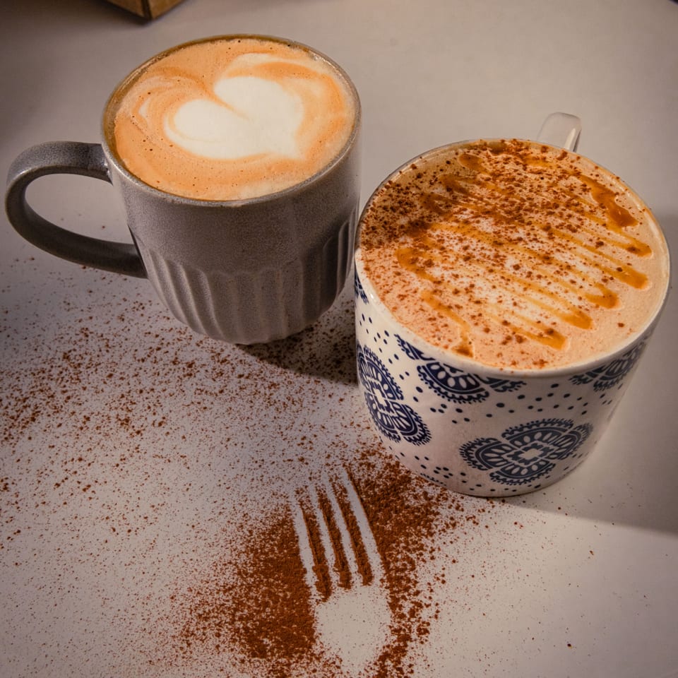 Two coffee mugs, one with latte art and the other with cinnamon and caramel drizzle, on a white surface.